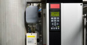 A lit display with a button control pad on a frequency converter in an industrial setting. The screen reads "0 rpm".