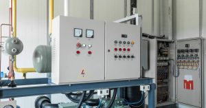 An industrial control panel in a control room. The panel has a hazard label and several rows of buttons.